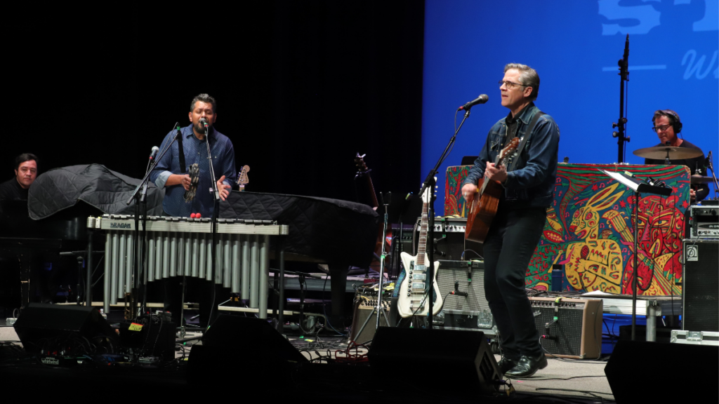 From left to right, a man plays the piano, a man plays the marimba, a man plays the guitar and sings, and a man plays the drums.