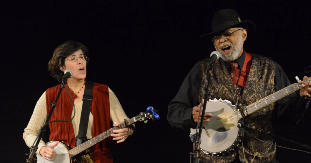 A woman and a man stand next to each other, both playing banjos and singing into microphones.