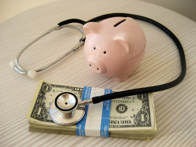 A piggy bank is shown with a stethoscope wrapped around it resting on a stack of dollars.