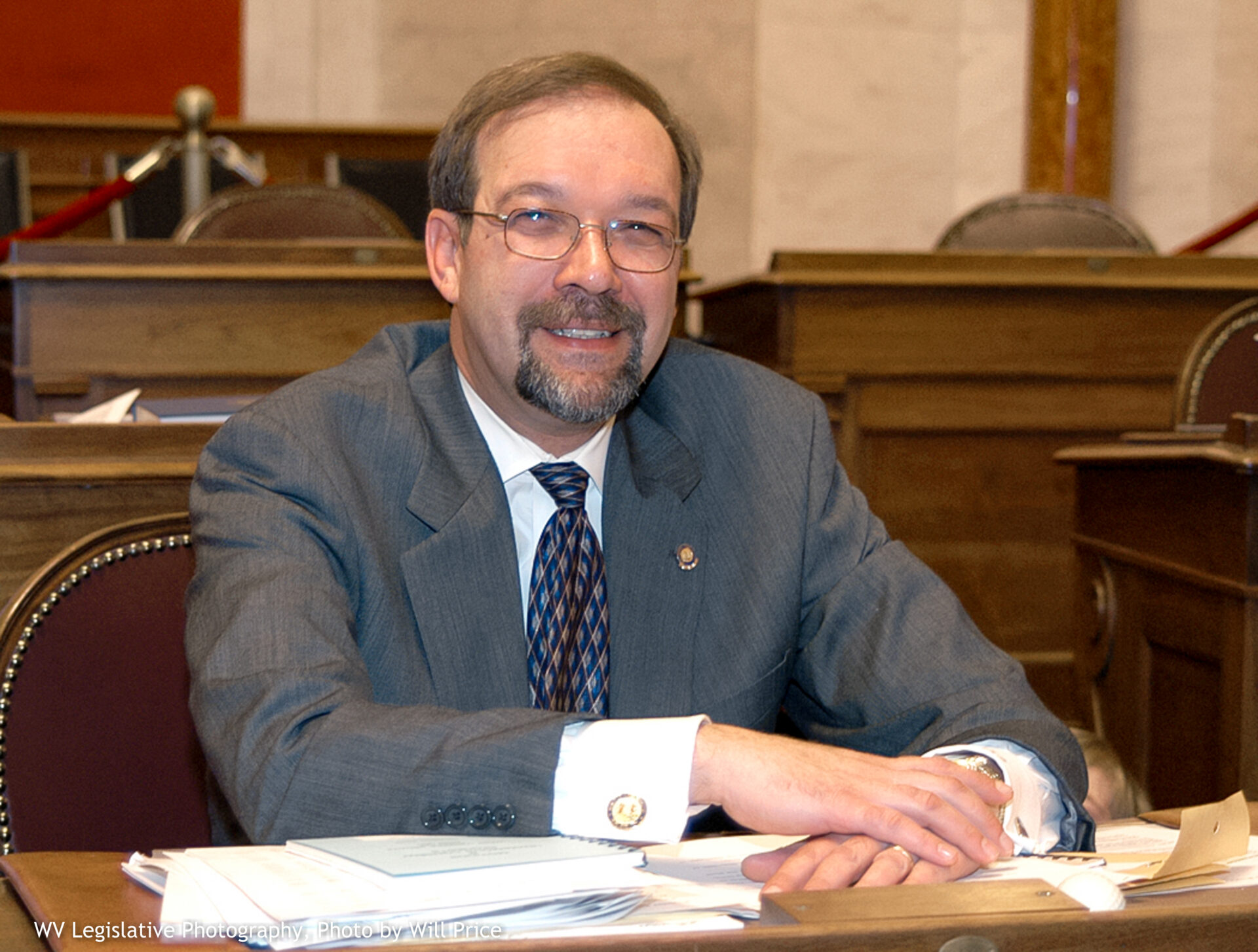 White man in a gray suit sits at a desk facing into the camera.