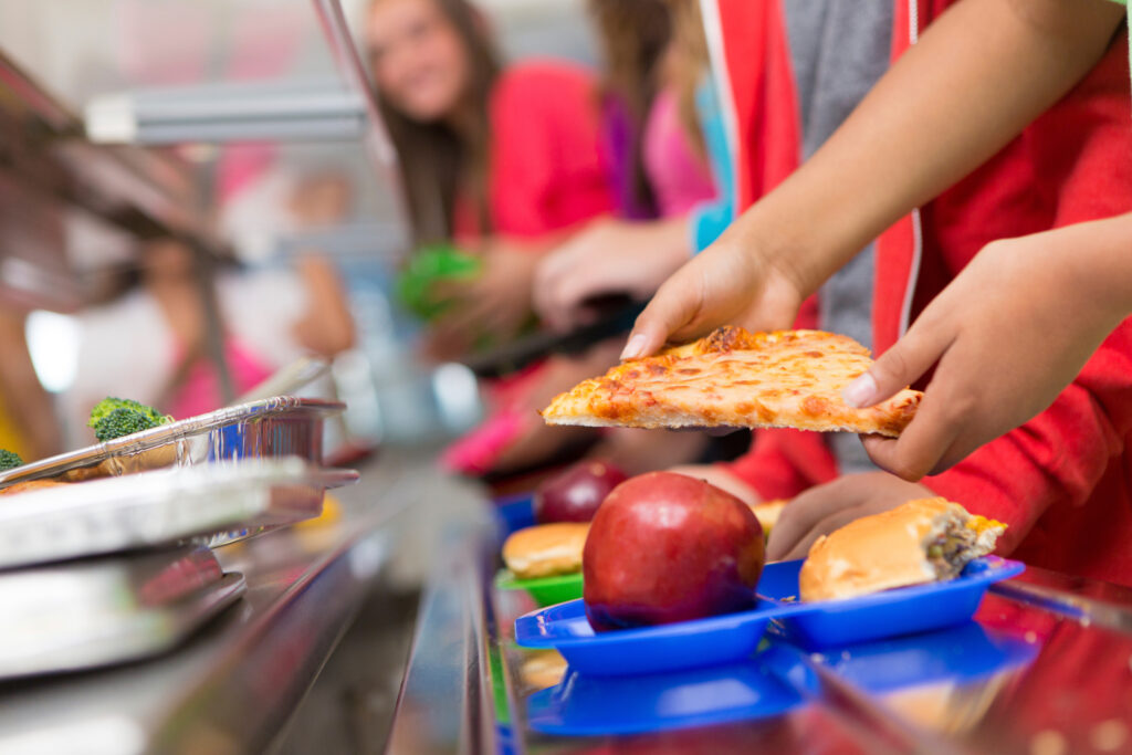 Focus on the foreground shows a hand placing a slice of pizza onto a school cafeteria tray. In the background, out of focus, students can be seen making their way down a counter.