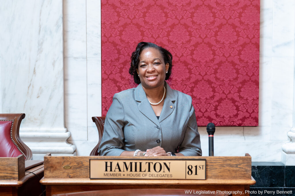 Del. Anitra Hamilton sits at her desk in the West Virginia House of Delegates after her swearing in ceremony. She is wearing a grey blazer and smiling, with a nameplate that reads "Hamilton, 81st." Behind her is a red pattern over white marble.