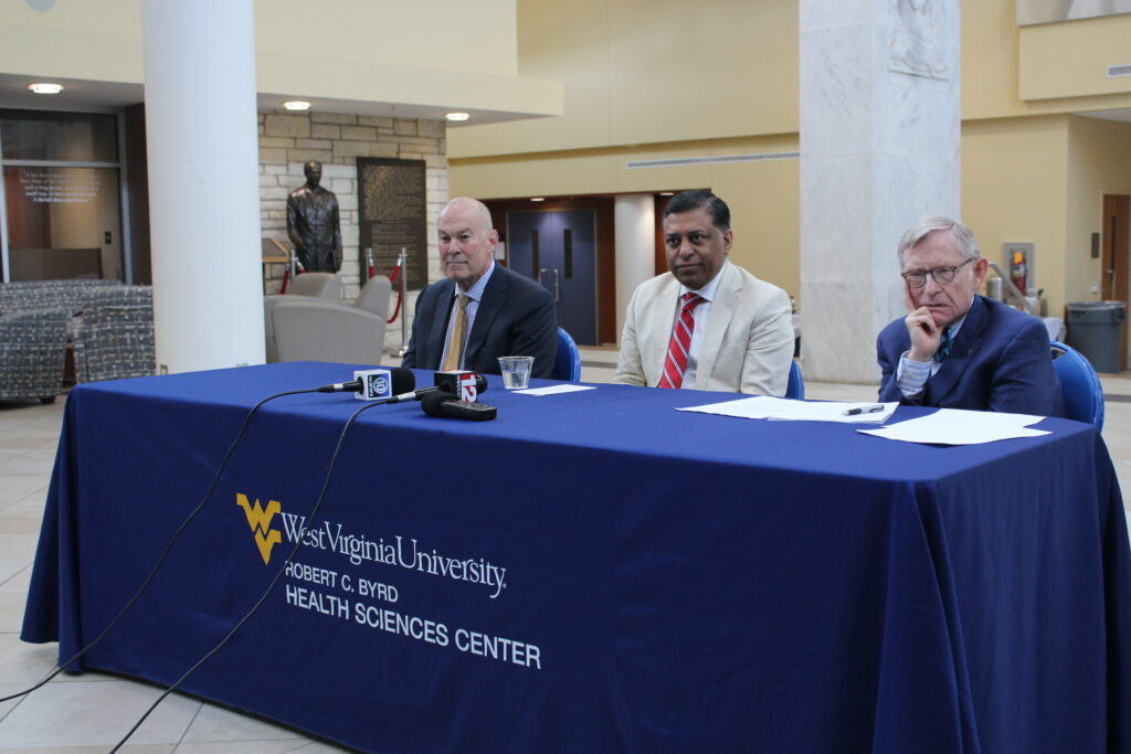 Three men sit at a table with a blue cloth draped over it. On the front of the cloth are the words "West Virginia University" Robert C. Byrd Health Sciences Center" There are two corded microphones on the table, as well as a bronze statue in the background
