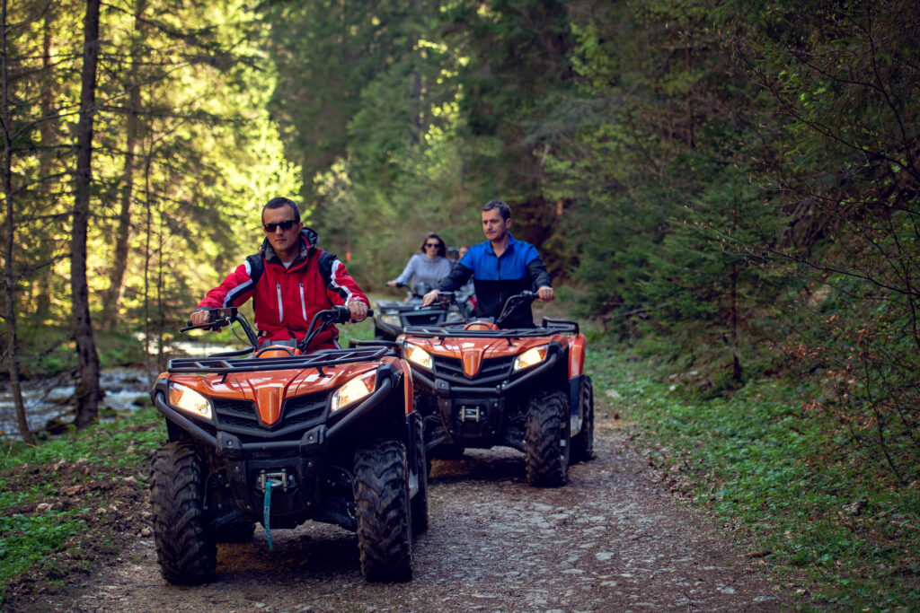 Three people riding ATVs . Two men, one woman. The man in the front is wearing a red jacket and sunglasses. The man behind him is in blue jacket and the woman in the rear is in a grey shirt.