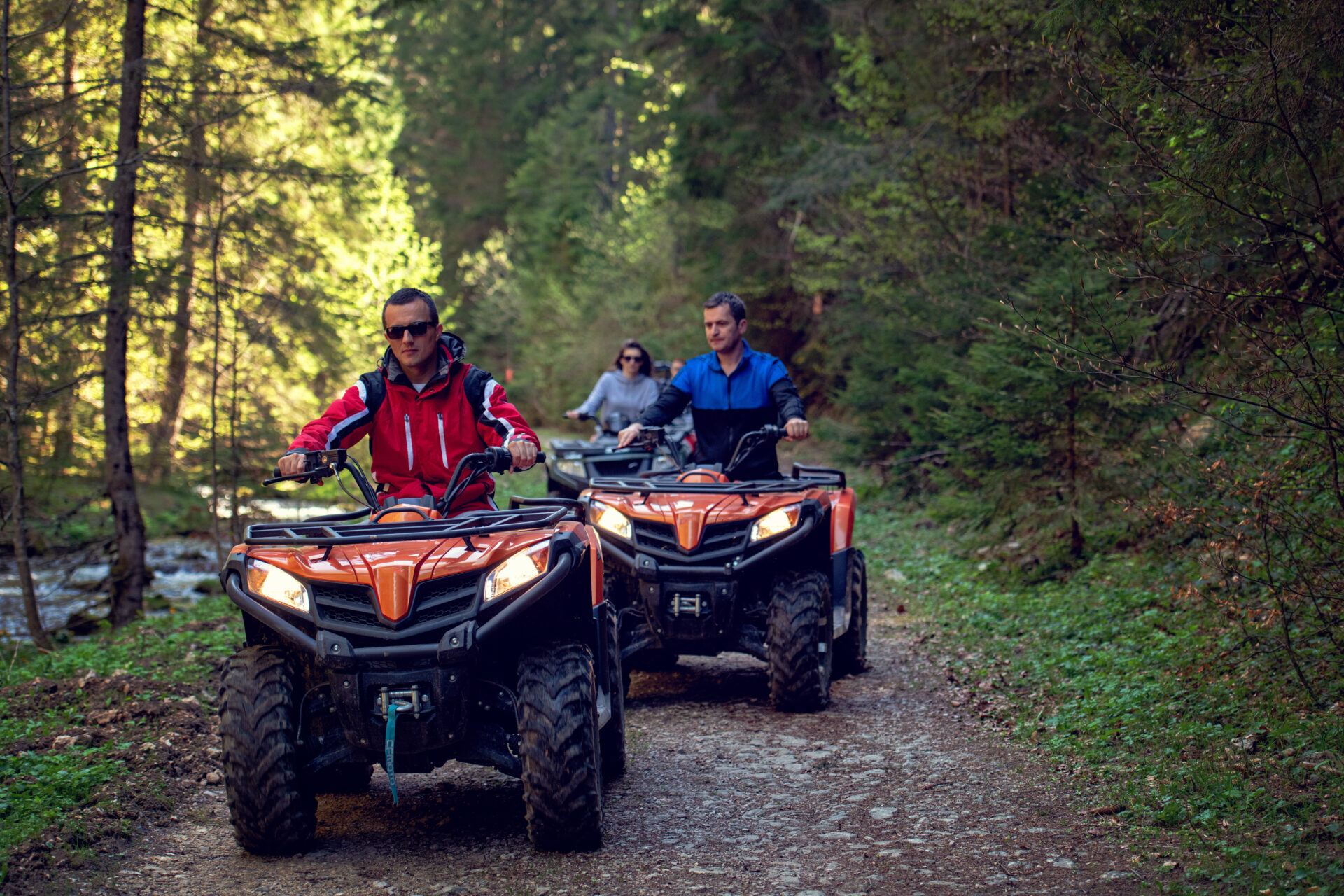 Three people riding ATVs . Two men, one woman. The man in the front is wearing a red jacket and sunglasses. The man behind him is in blue jacket and the woman in the rear is in a grey shirt.