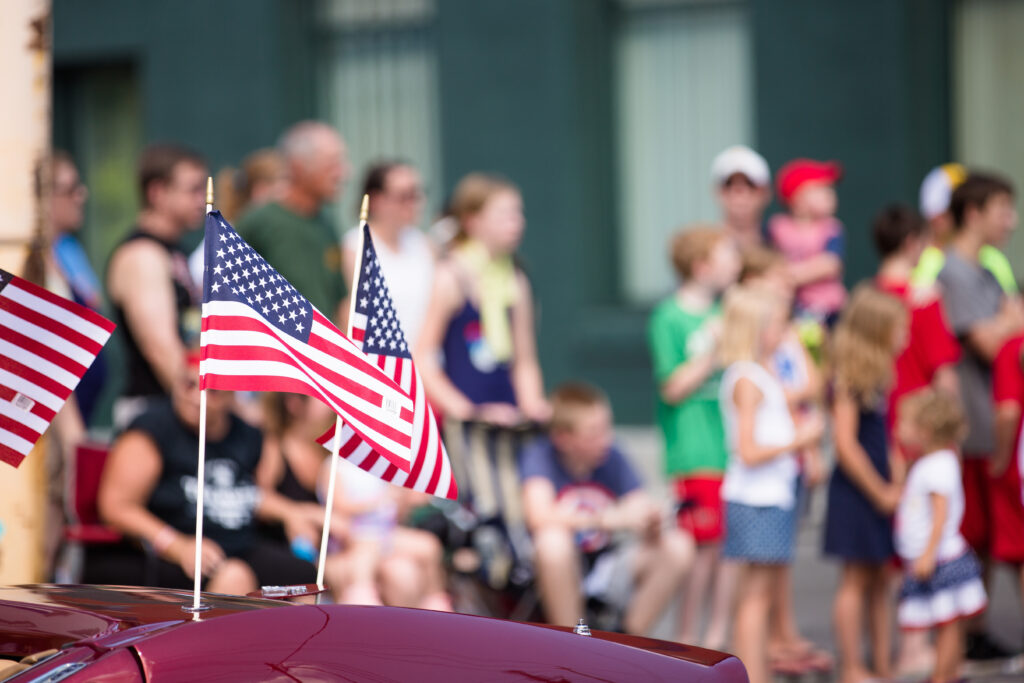 American flags are seen on the hood of a car. In the distance are people standing watching a Fourth of July parade.