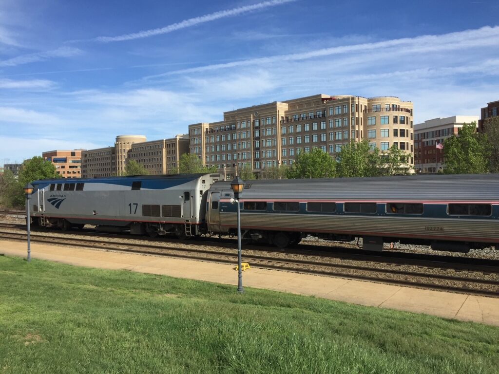 An Amtrak train stops in Alexandria, Virginia in 2017. The train has a single locomotive and the train is bracketed by the station platform and the city skyline.