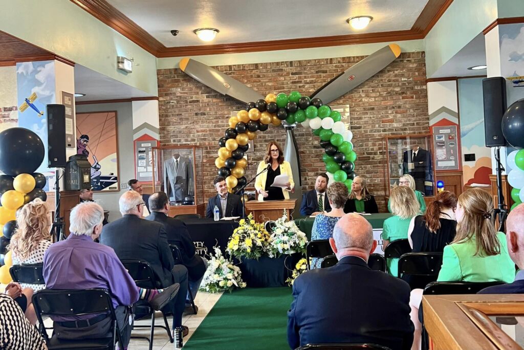 A room full of people listening to a woman dressed in yellow and black who is standing at a podium in front of a propeller and colorful balloons. The woman is announcing the opening of a new flight school at Wheeling-Ohio airport.