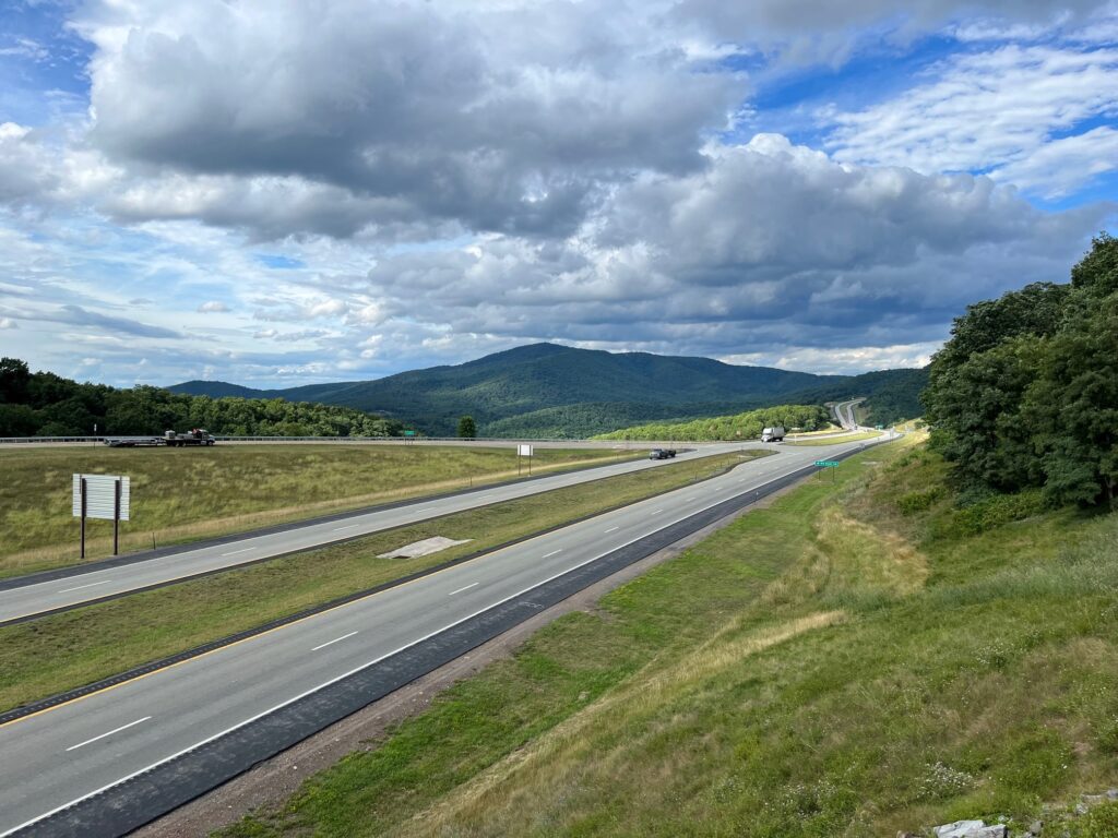 A four-lane highway, viewed from an overpass, winds through the mountains with clouds and sun.