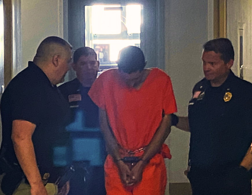 Man in prison orange jumpsuit flanked by police