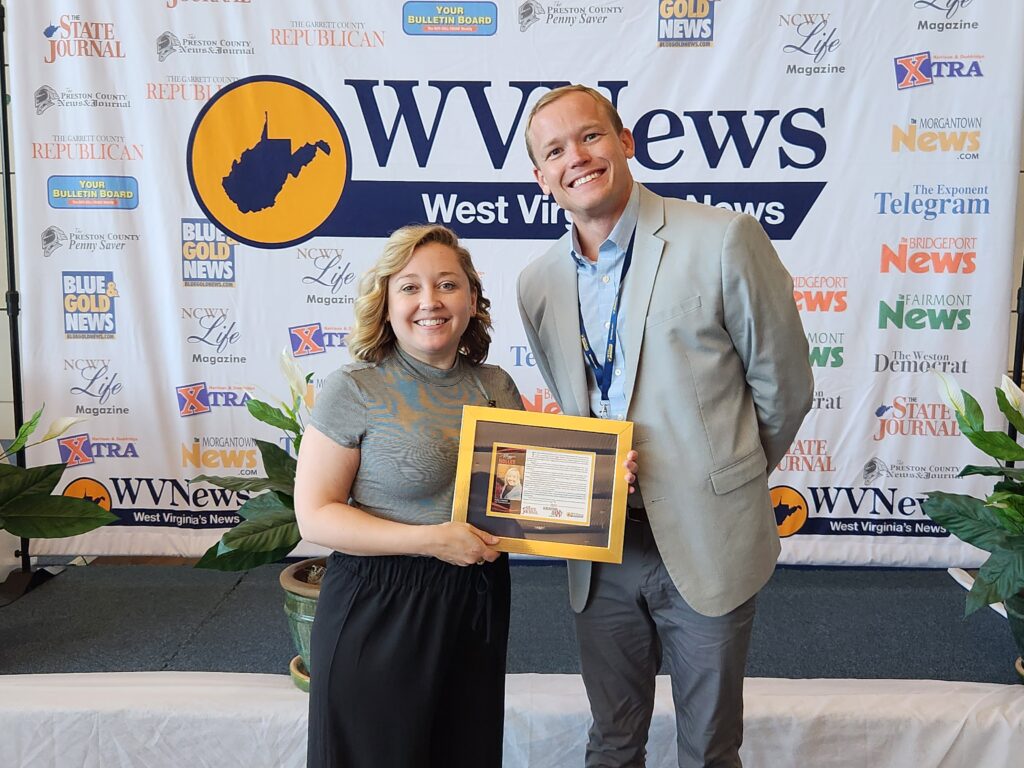 A woman and man stand side-by-side, smiling. The woman holds an award. Behind them is a backdrop for photos with "WVNews" in large letters.