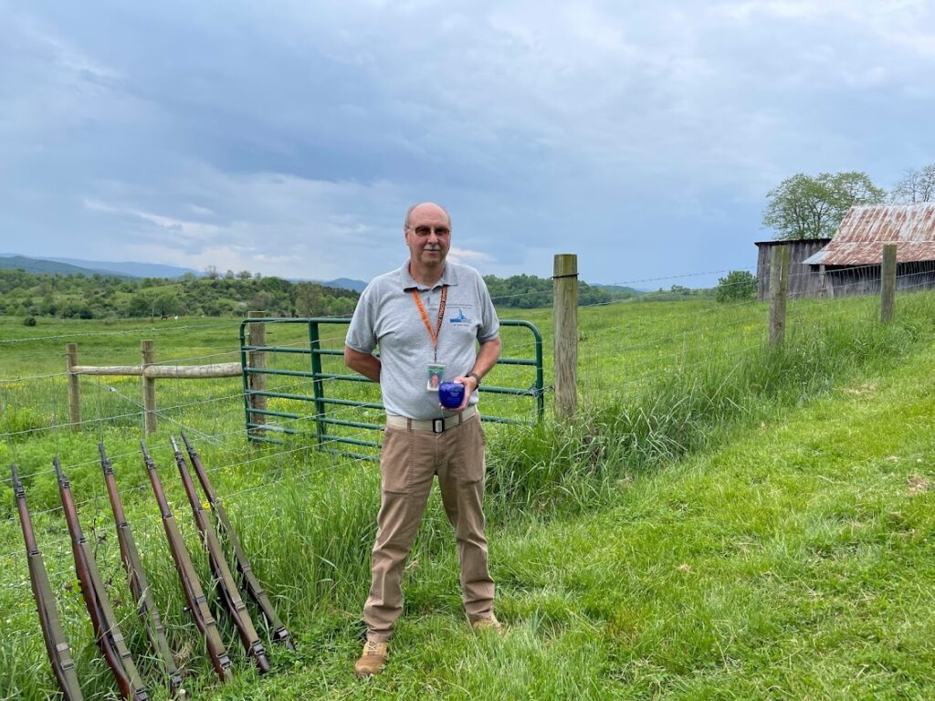 Mike Wiley stands in a field with a wooden fence in the background, blue, cloudy sky, and rolling hills in the far background.