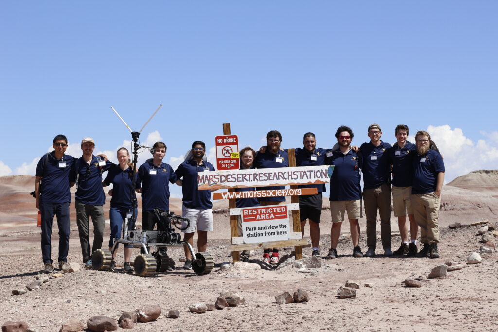 A group of West Virginia University students all wearing dark blue polos stand behind a set of signs that read, from top to bottom, "No Drone" "MarsDesert.ResearchStation" "www.marssociety.org" "Restricted Area-Park and View the Station from Here" in a dry landscape under a clear blue sky.