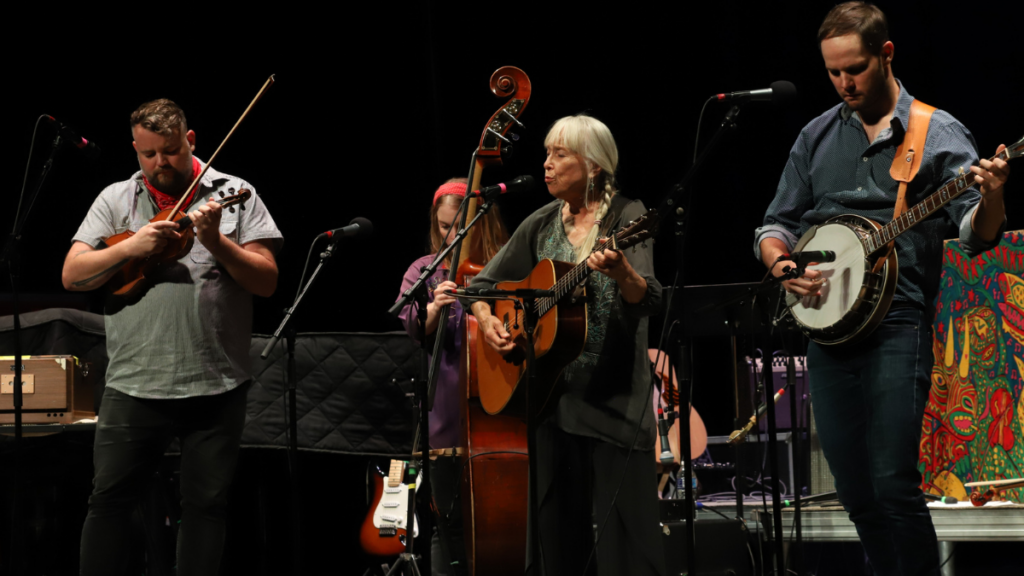 Four musicians perform bluegrass music on a stage with a fiddle, upright base, guitar and banjo.