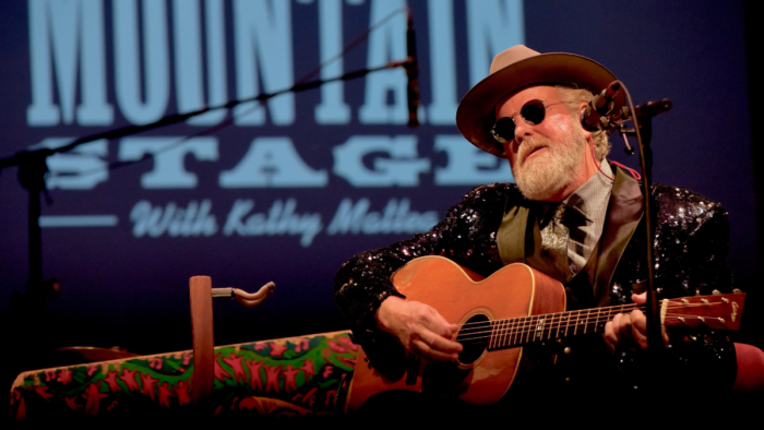 A man with grey beard, hat, and sunglasses sings and plays the guitar.