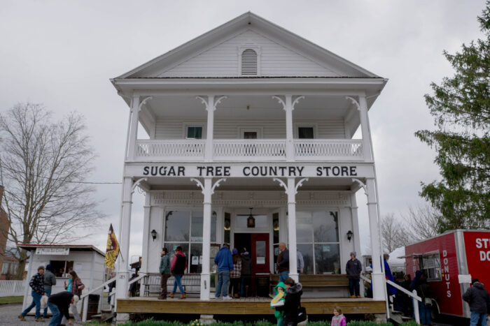 A large white building, colonial style is seen in the center of the image. People are walking up on the porch and along the road in front of it. The words "Sugar Tree Country Store" are seen on the building.