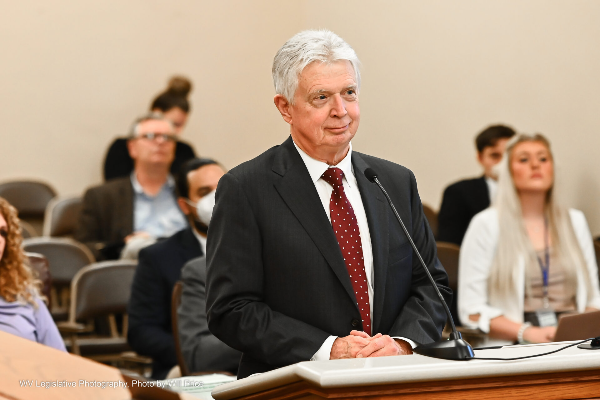 Superintendent David Roach stands at a lectern while speaking to the Senate Committee on Education in early 2023. He wears a black suit over a white shirt and red tie. Behind him is arrayed the gallery of the meeting chambers.