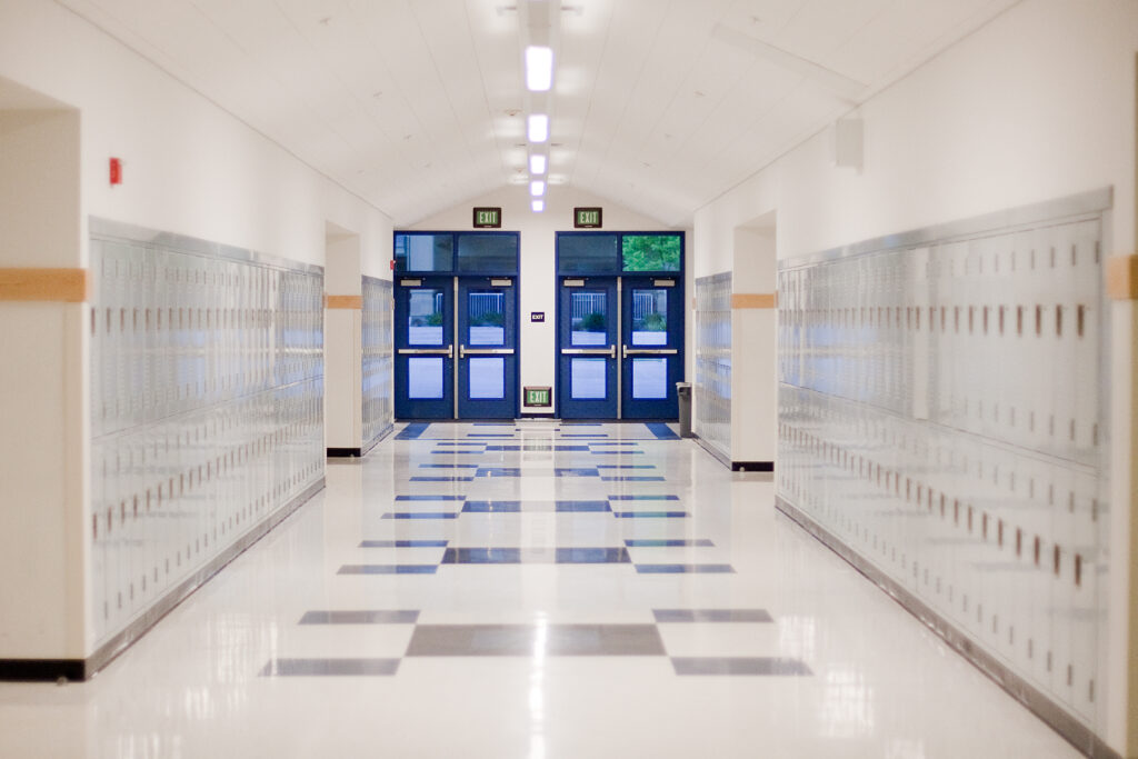 An empty school hallway is largely white and well-lit, with bright blue doors at the end of the hallway.
