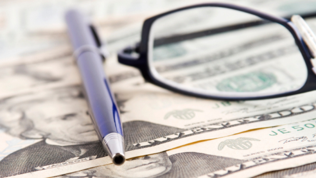 Glasses and a blue pen are seen on top of several twenty dollar bills.