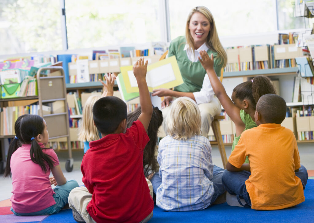 A group of young children, many with a hand raised, sit on the floor in front of a smiling, blonde woman wearing a light green sweater and holding a picture book in front of the group. The scene is set in front of shelves of books in the background.