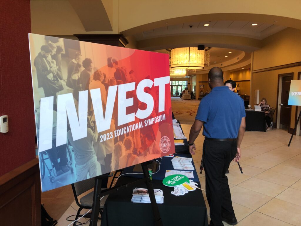 A colorful background contrasts the white lettering "Invest 2023 Educational Symposium" on a sign in a hallway. A man in a blue shirt and black pants stands next to the sign with his back to the camera in a hallway that extends away from the camera.
