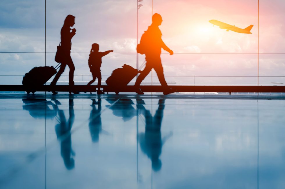 A family walking with their hand luggage through an airport with a plane observed taking off in the distance during sunset.