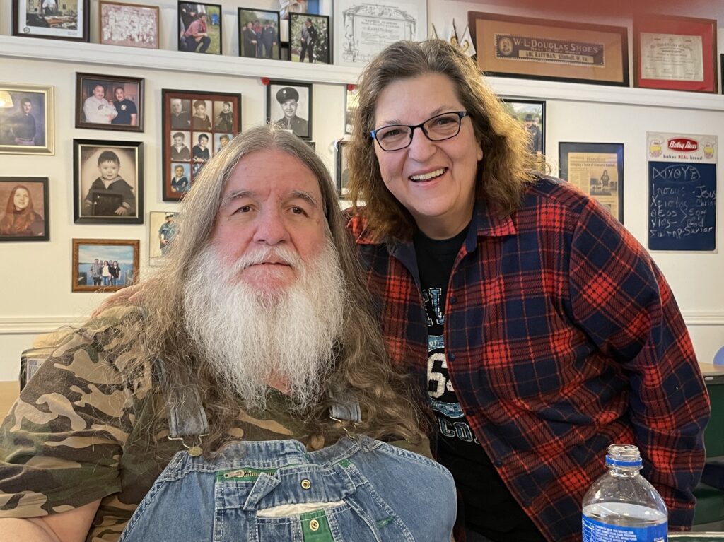 Two people stand next to each other. Both smiling. One man with a white beard in overalls and a woman with brown hair and glasses wearing a flannel shirt.