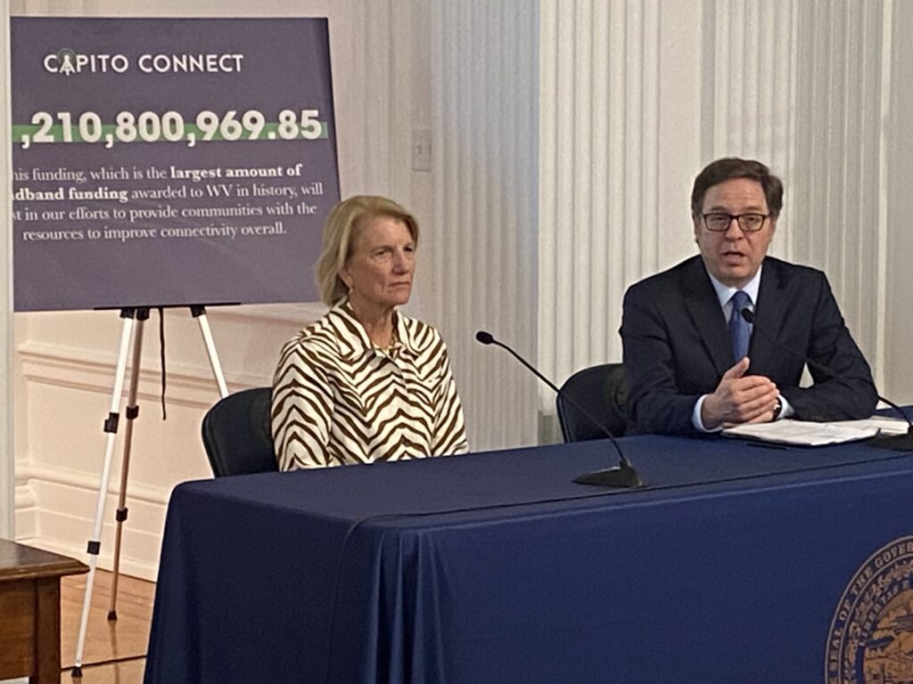 Blond, petite woman sitting next to man in suit and wearing glasses.