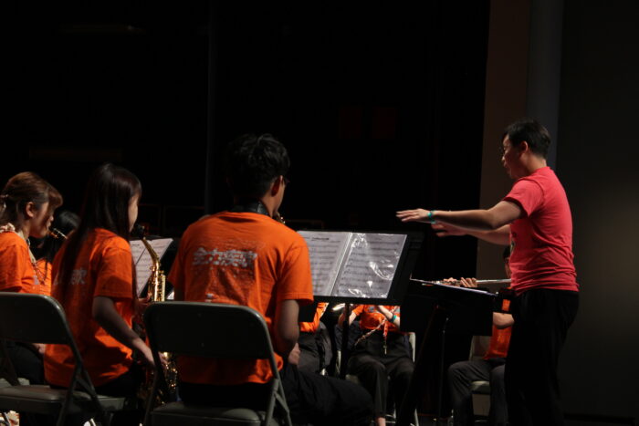 Musicians dressed in orange shirts play saxophones while seated in front of sheet music on stands. A man in a red shirt on the right of frame gestures at the musicians.