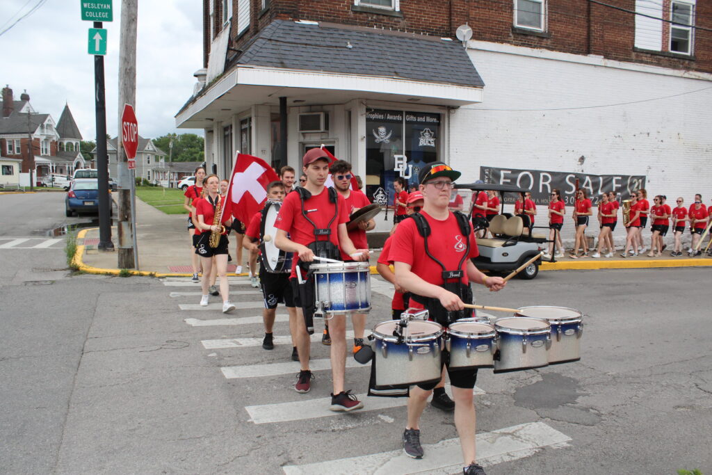 A drummer dressed in a red shirt and a black cap leads a group of young musicians on a march across a street in front of a whitewashed brick wall.