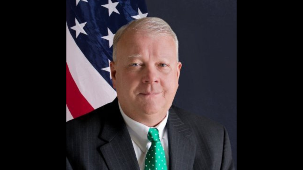 Man with white hair and in a formal suit. Headshot photo in front of the American flag.