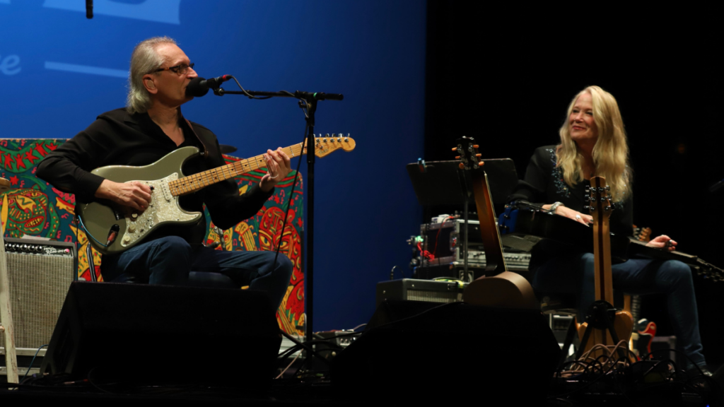 A man plays the guitar sitting down while singing and looking at a woman playing the slide guitar to his left. She is looking back at him smiling.