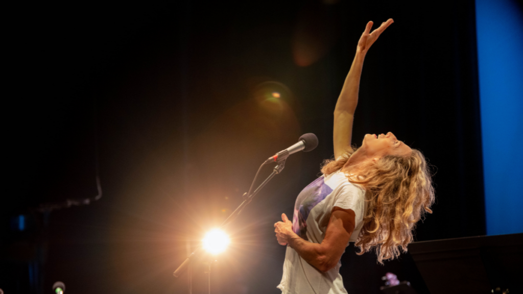 A woman with sandy brown curly hair leans back looking up with her right hand raised in the air in front of a microphone.