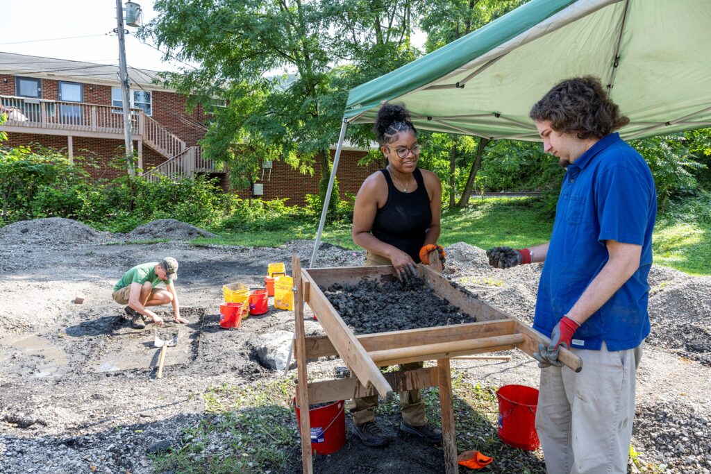 Two students, a woman and man, stand over a soil shifter filled with dirt. The woman is in a black tank top, while the man is in a blue shirt. They stand under a tent. A brick house and green trees are seen in the background.