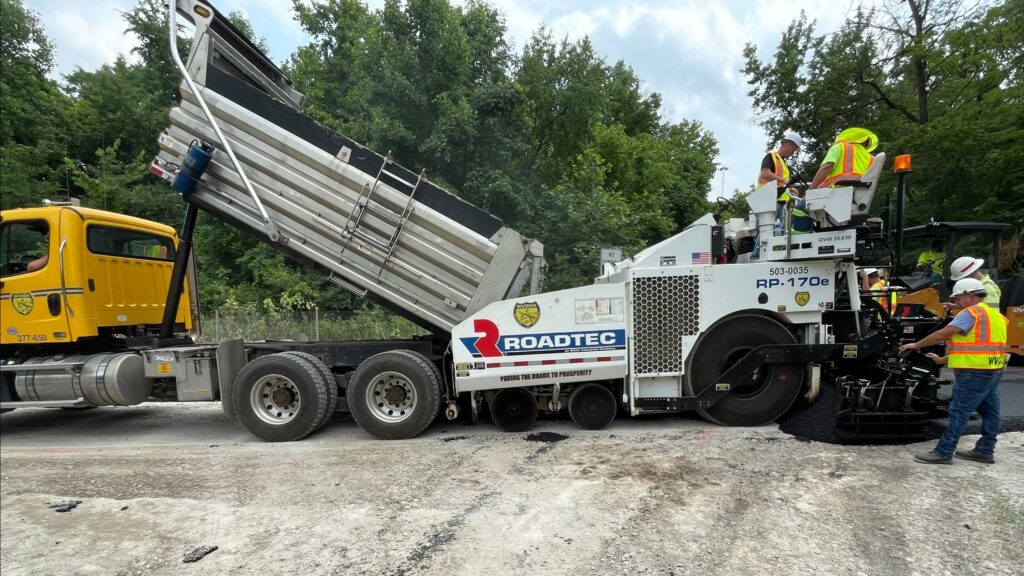 A yellow truck dumps materials into a white pavement machine. The machine has the word "Roadtec" written across it.