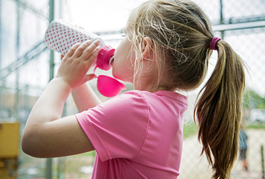A young girl is seen drinking from a pink water bottle on a hot summer day.