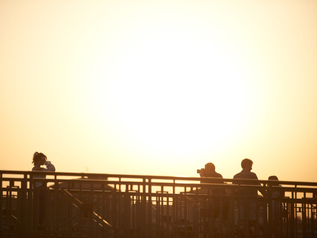 A bright yellow sun that dominates the top of the frame with several people standing at the bottom of the frame silhouetted behind a railing.