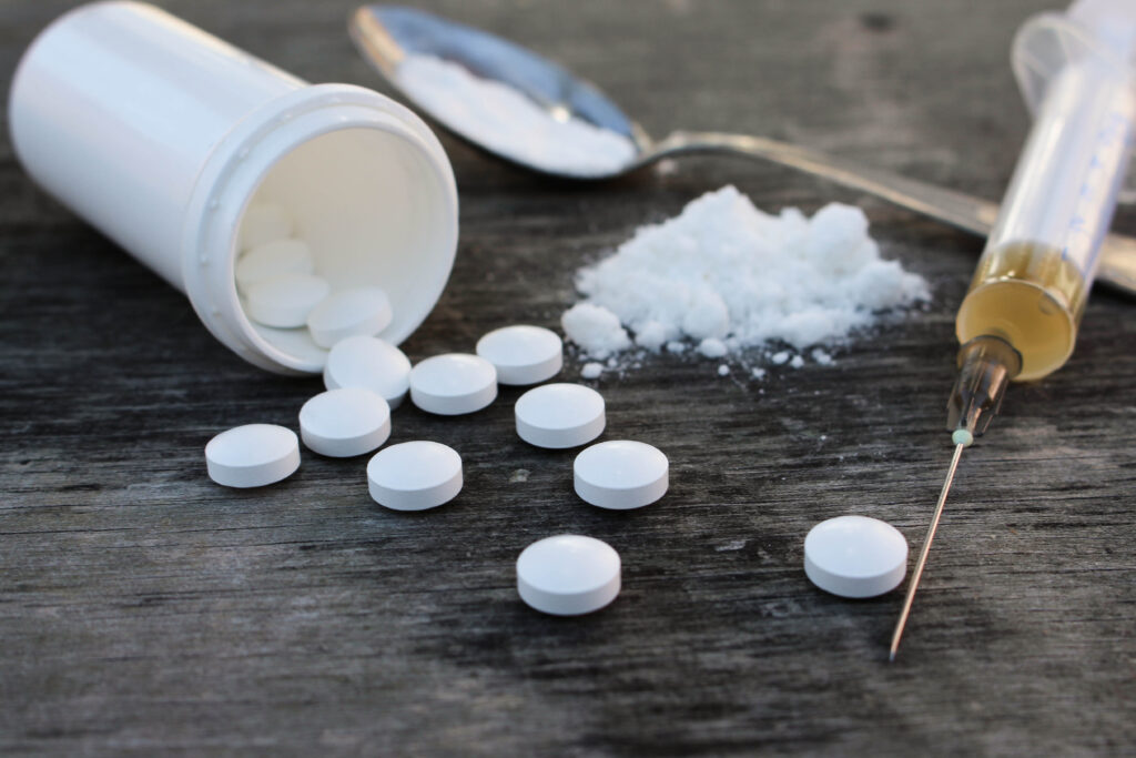 A close up of white pills, a syringe, and a spoon with a white substance are seen on a wooden table. This is a stock image of opioids.