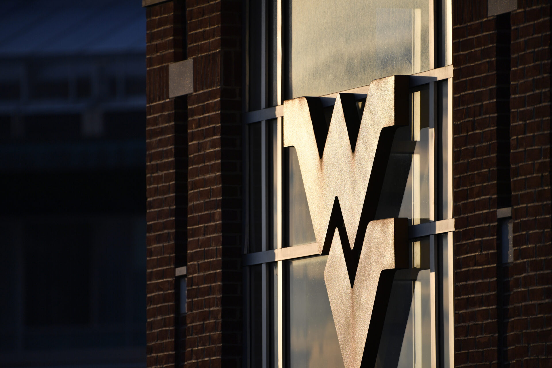 A West Virginia University logo, known as the "Flying WV" can be seen on the facade of a building. The letters are lit up by sunlight.