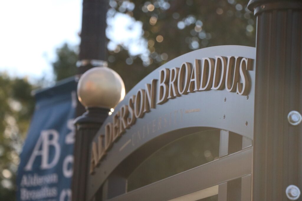 A metal arch between lamp posts reads "Alderson Broaddus" with the word "University" just visible below. In the background, out of focus is a blue banner hanging off a different pole with the letters "AB" visible in white script. The entire image is suffused with light from just off frame shining directly into the lense.