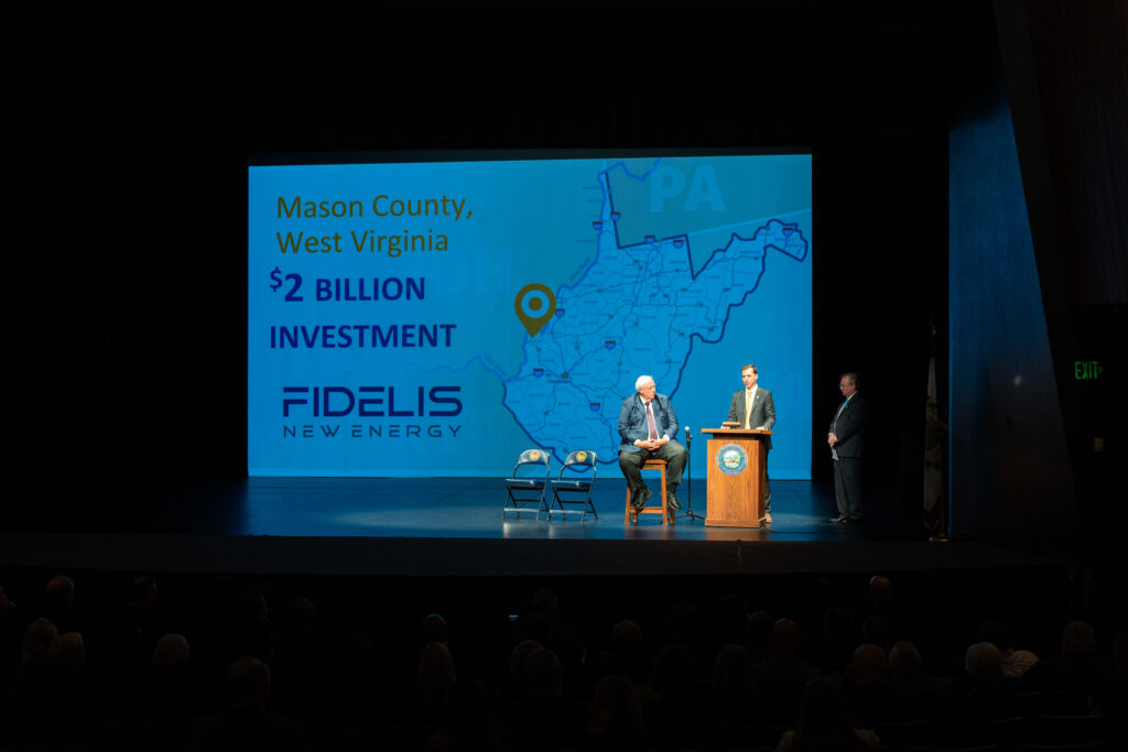 Gov. Jim Justice sits and two men stand on stage in front of a projection that reads "Mason County, WV $2 Billion Investment Fidelis New Energy" next to a map of the state with a pin in Mason County.