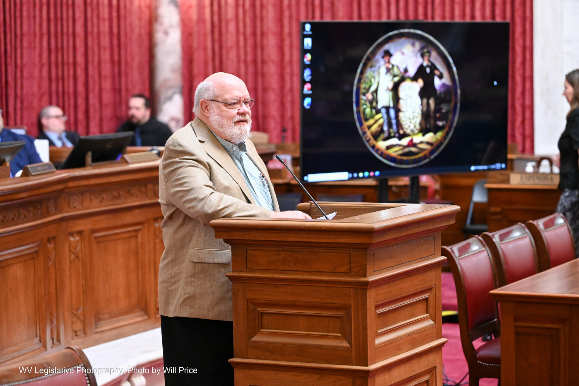 A man with white hair and beard stands at a lectern in front of empty desks. Behind him to the right can be seen a flatscreen TV displaying a stylized state seal. On his other side can be seen a podium and red curtains against the wall.