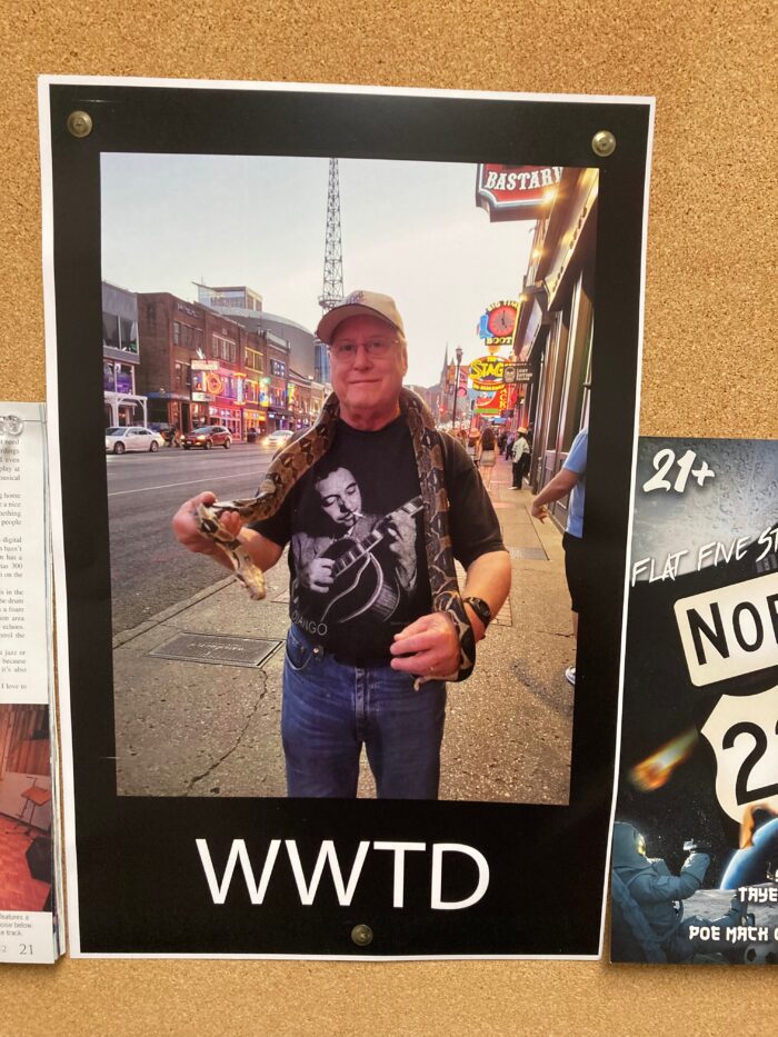 A photograph is shown on a pin board. An older man in the photo has a large snake draped over his shoulders. He wears a ball cap and glasses.