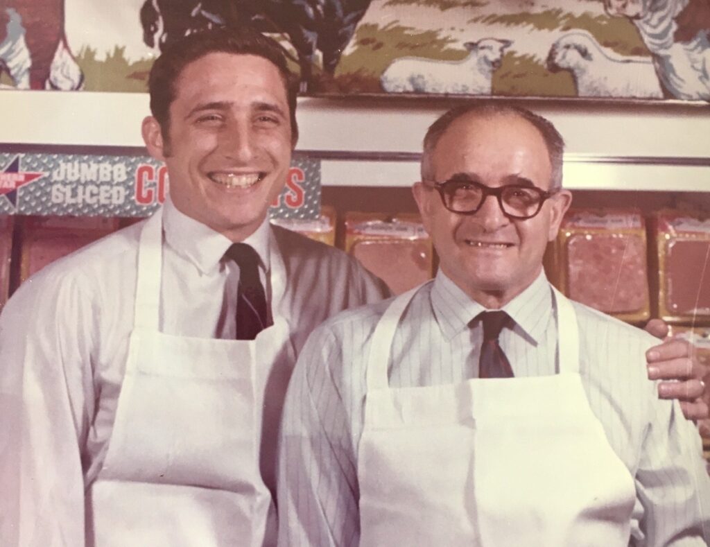 Two men stand next to each other in white aprons smiling for the camera. The men are also in business casual attire - button up shirts with ties.