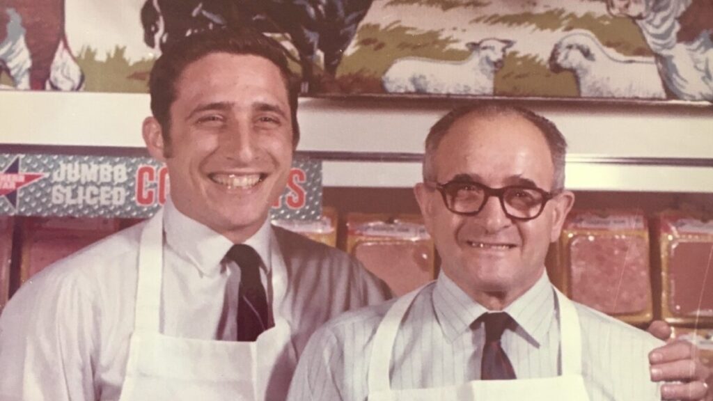 Two men stand next to each other in white aprons smiling for the camera. The men are also in business casual attire - button up shirts with ties.