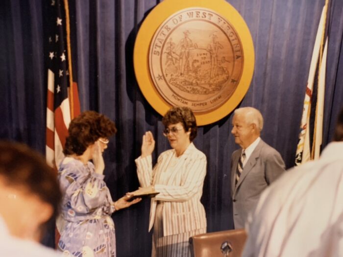 Donna Boley stands with left hand on bible and right hand in air. She is being sworn in to the Senate.  The man standing behind her is the governor who appointed her to the position. They are standing on a stage with the round West Virginia emblem behind them. 