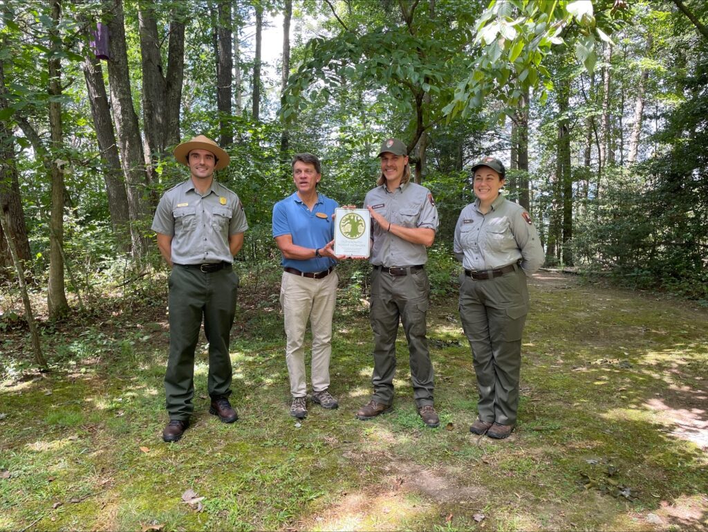 Three park rangers, Rasso, Mann, and Katie Kull stand with Kane from the Old Growth Forest Network for a photo. They are holding a plague and standing in front of a forested area.
