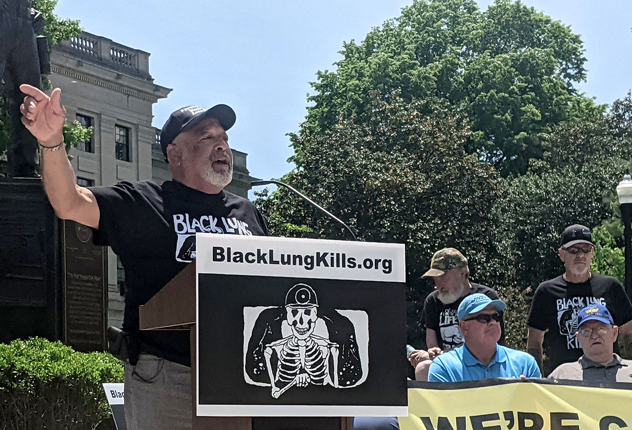 A gray bearded man wearing a hat and a black shirt waves at a crowd carrying signs in front of a granite building on a clear day.
