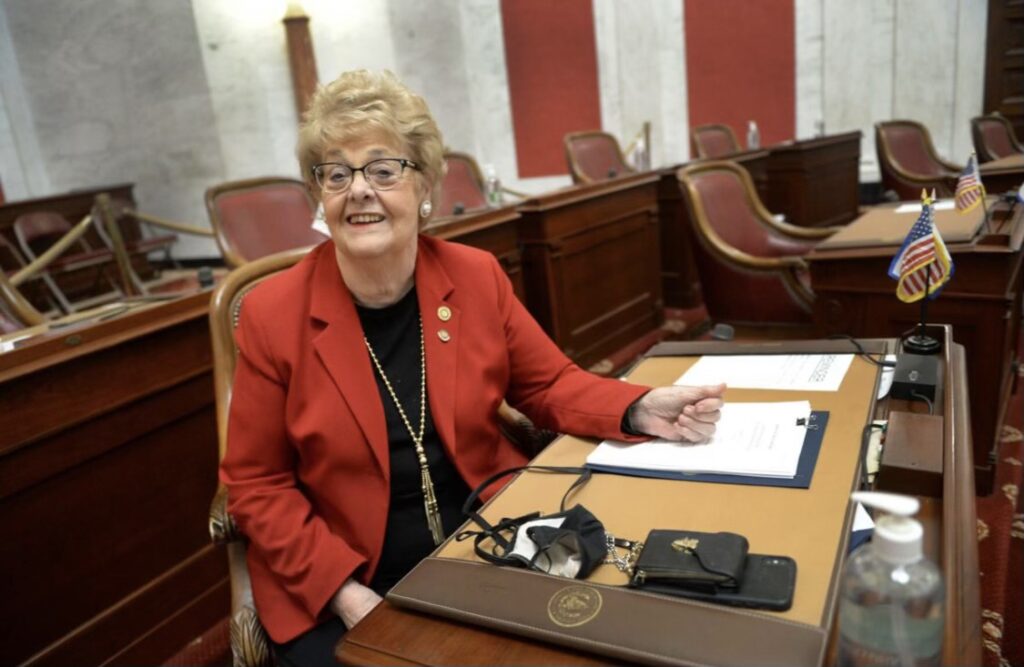 Senator Boley sits at a desk in the state senate wearing a bright red blazer, with gold accessories. She is looking at the camera smiling.