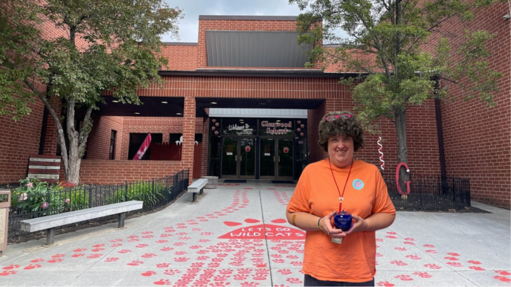 A woman in an orange shirt, holding a blue glass apple smiles for the camera. She stands in front of the front doors of a school.